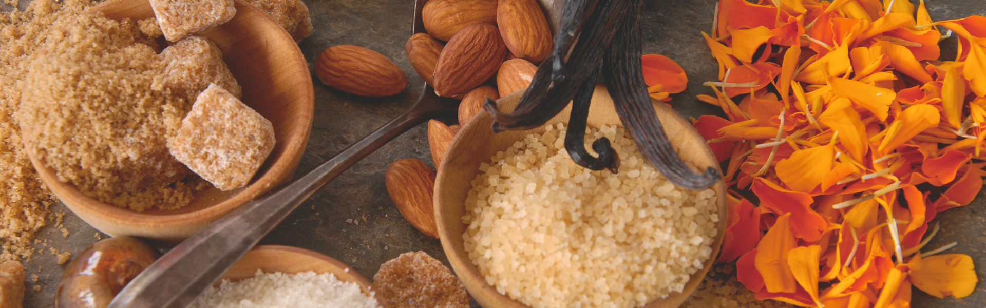Spices and herbs including almonds, flowers, and a bowl of sugar on a textured surface.