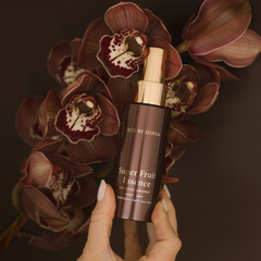 Bottle of 'Super Fruit Essence' by Sonya held in front of dark pink orchids.