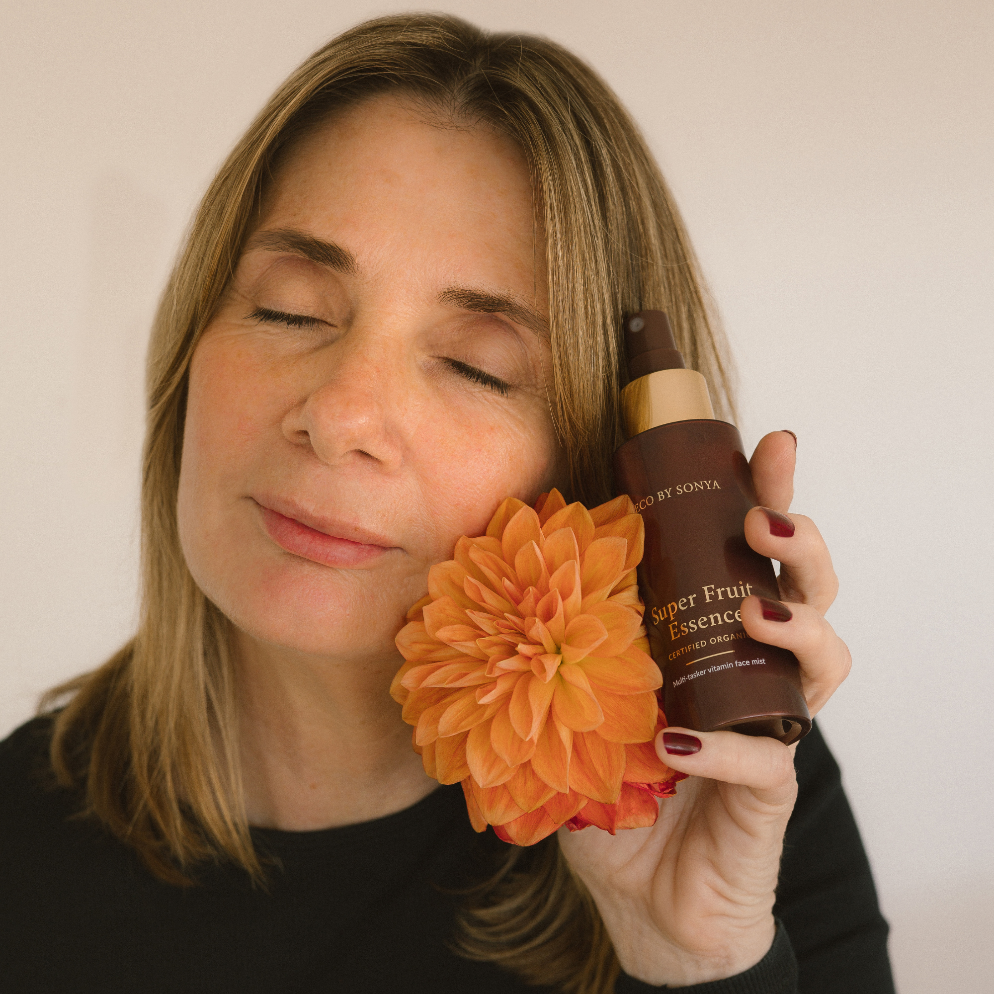 Woman holding a bottle of 'Super Fruit Essence' and an orange flower against a plain background