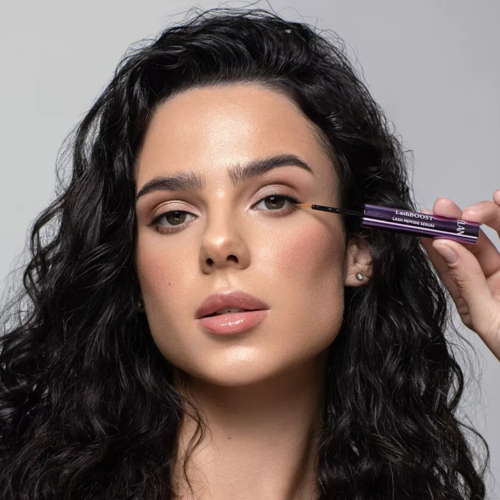 Woman applying mascara with a purple and white mascara wand against a gray background